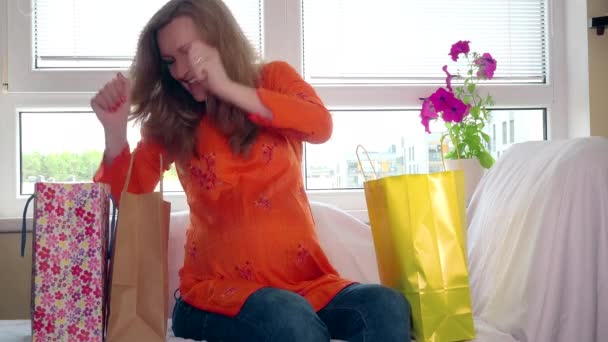 Mujer embarazada feliz después de ir de compras sentado en el sofá y mirar la ropa del bebé — Vídeos de Stock