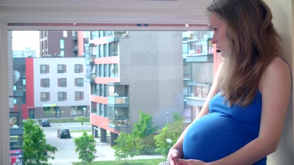 Грустная беременная женщина смотрит в окно и нежно ласкает своего ребенка шишку — стоковое видео