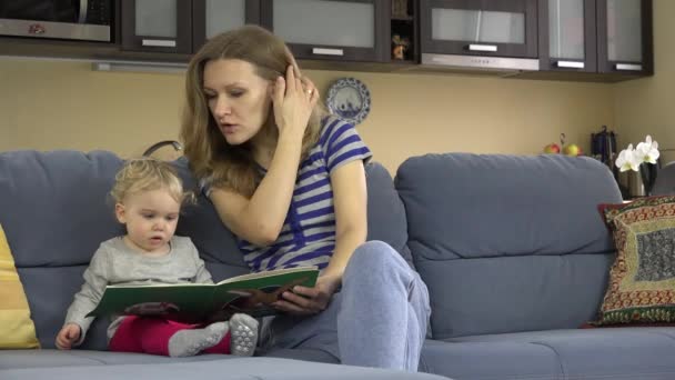 Mother read book with little baby daughter on sofa in room. 4K — Stock Video