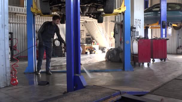 Arbetaren kille rent smutsiga golvet under hängande bil i verkstad. — Stockvideo