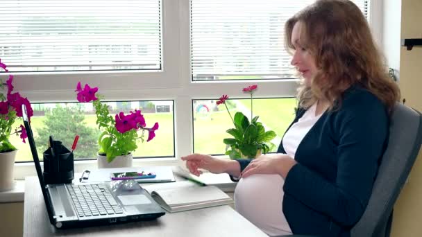 Уставшая беременная девушка сидит рядом с компьютером и гладит большой животик — стоковое видео