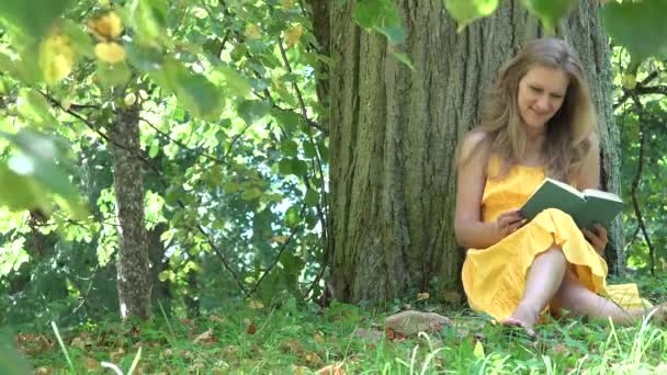 Niedliche Frau in gelbem Kleid sitzen unter alten Baum genießen Roman Buch im Sommerpark lesen. 4k — Stockvideo