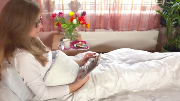 Женщина сидит в постели за планшетным компьютером. Завтрак и цветы на шкафу — стоковое видео