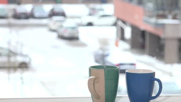 Две чашки кофе чай горячий напиток стоять на подоконнике и снежинки снег падает — стоковое видео