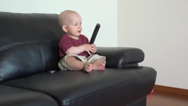 Lustige Jungen drücken Tasten der Fernbedienung sitzt auf schwarzem Sofa — Stockvideo