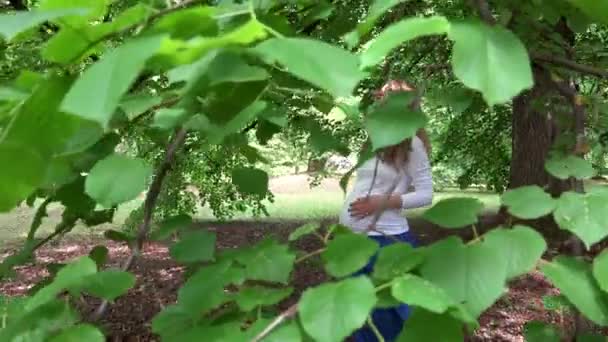 Улыбающаяся беременная женщина под ветвями старого дерева. Листья движутся на ветру — стоковое видео