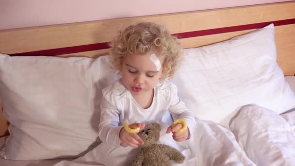 Дівчина Тоддлер з покритою раною на голові їсть кукурудзяне печиво, сидячи на ліжку — стокове відео