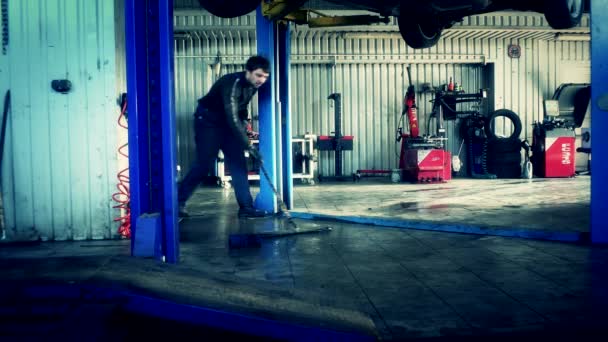 Worker man clean dirty floor under hanging car in repair garage. — Stockvideo