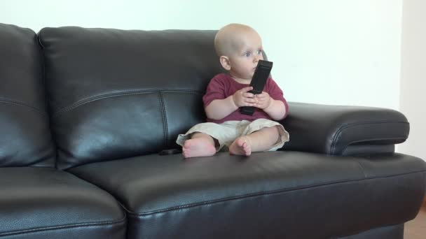 Carino bambino seduto sul divano nero con telecomando a casa — Video Stock