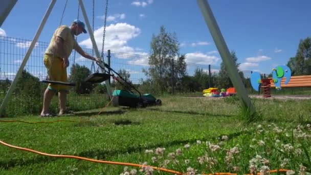 Młody człowiek kosi trawnik z placem zabaw dla dzieci. Ruch Gimbal — Wideo stockowe