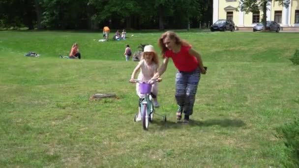 Madre ayuda a su hija de 4 años a andar en bicicleta por el prado del parque — Vídeo de stock