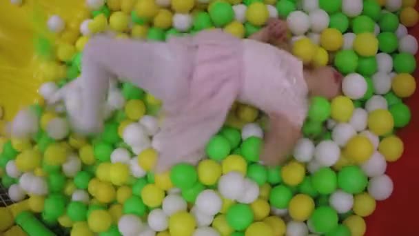 Hiperaktív lány rózsaszín ruhában játszanak a játszótéren tele műanyag színes golyókat — Stock videók