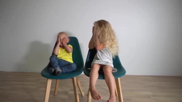 Niños juguetones hermano y hermana se sientan en sillas cubren los ojos mirando a la cámara . — Vídeo de stock