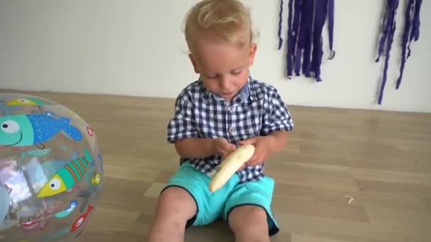 Liten pojke på två år som sitter på golvet och äter banan. Gimbal rörelse — Stockvideo