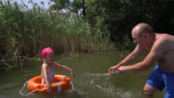 Vater und Tochter spritzen Wasser. Mädchen mit Badeanzug und Kopftuch im Rettungsring — Stockvideo