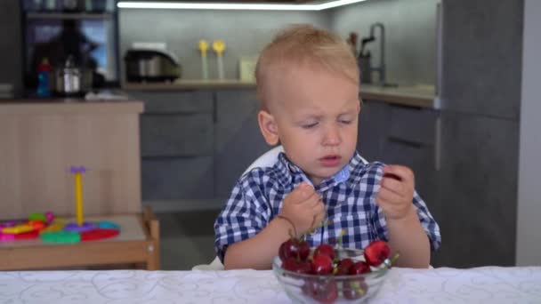 Portret niewinnego chłopca jedząc czereśnie w domu przy stole. Gimbal Motion shot. — Wideo stockowe
