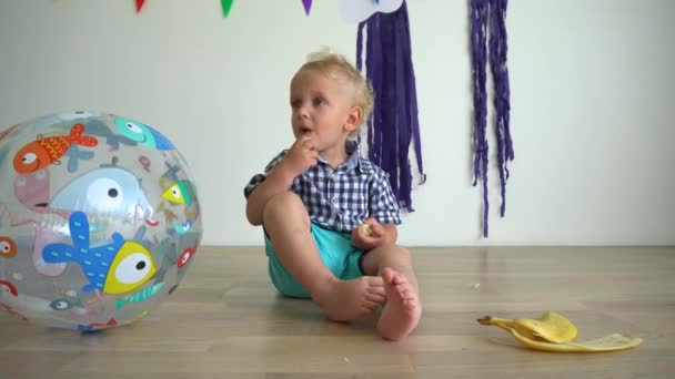 Aantrekkelijke jongen 2 jaar oud eet een banaan. Het kind zit op de verdieping. Gimbal Motion — Stockvideo