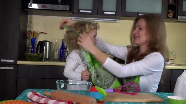 Lächelnde Frau bindet Schürze auf kleines Mädchen am Küchentisch. glückliche Familienzeit — Stockvideo