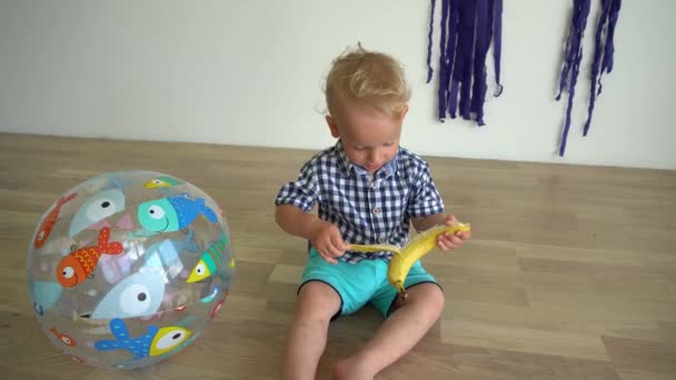 Niño pelando plátano sentado en el suelo de madera en casa. Movimiento del cardán — Vídeos de Stock