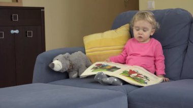 Küçük meraklı kız görünümlü görüntüler kitap ve oyuncak ayı mavi kanepede oturan