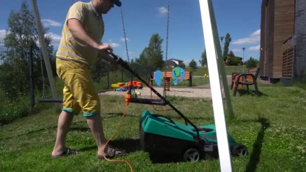 Der junge Mann arbeitet auf einem bunten Kinderspielplatz. Mann mähte Gras zwischen Schaukel — Stockvideo