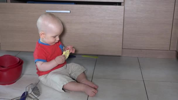 Голодный мальчик ест еду, сидя в беспорядке на полу на кухне — стоковое видео