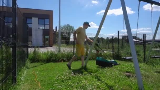 Мужчина стриг траву газонокосилкой во дворе дома рядом с детской площадкой. Гимбальное движение — стоковое видео