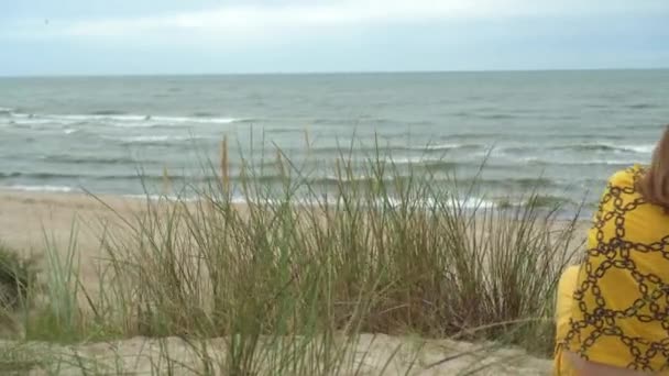 Jeune femme assise dans les dunes parmi l'herbe haute et versant du sable entre les mains — Video