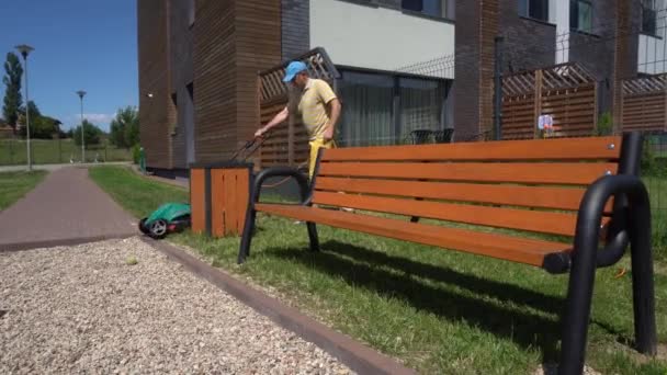 Trädgårdsmästare man klippa gräsmatta med elektrisk maskin nära träbänk. Gimbal rörelse — Stockvideo