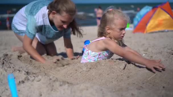 Mother and daughter have fun on seaside. Woman digging little girl deep in sand — Stock Video