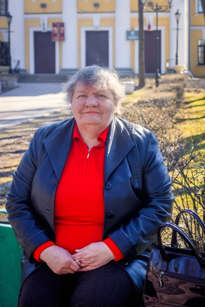 Oudere vrouw op een bankje in de stad. Grootmoeder in een rode trui en lederen jas. — Stockfoto