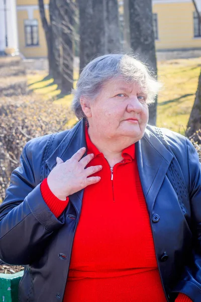 Oudere vrouw op een bankje in de stad. Grootmoeder in een rode trui en lederen jas. — Stockfoto