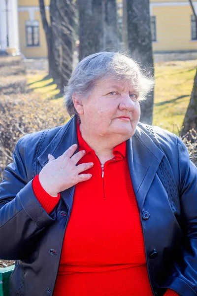 Oudere vrouw op een bankje in de stad. Grootmoeder in een rode trui en lederen jas. — Stockfoto