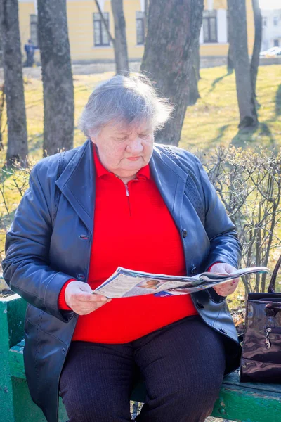 坐在长凳上看报纸的老年妇女。穿着红色毛衣和皮夹克的祖母。祖母读报纸 — 图库照片