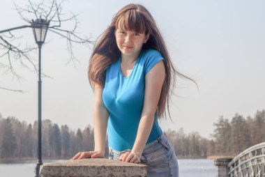 Kotlu ve tişörtlü genç bir kadın baharda göl kenarında poz veriyor.