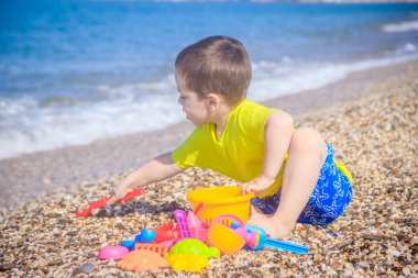 Küçük çocuk deniz arka planda renkli oyuncaklar ile çakıl plajda oynuyor 