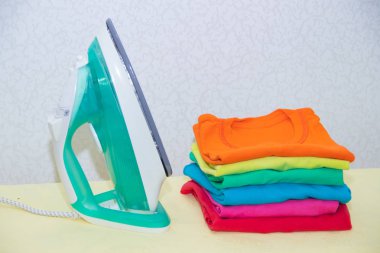 Demir ve bebek kıyafetleri. Ütü tahtasında renkli kıyafetler. Parlak tişörtler. Bir tahta üzerinde ütülenmiş renkli bebek iç çamaşırı. . Ütü tahtası. Bir yığın şey.