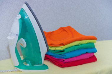 Demir ve bebek kıyafetleri. Ütü tahtasında renkli kıyafetler. Parlak tişörtler. Bir tahta üzerinde ütülenmiş renkli bebek iç çamaşırı. . Ütü tahtası. Bir yığın şey.