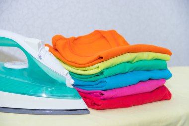 Demir ve bebek kıyafetleri. Ütü tahtasında renkli kıyafetler. Parlak tişörtler. Bir tahta üzerinde ütülenmiş renkli bebek iç çamaşırı. . Ütü tahtası. Bir yığın şey.