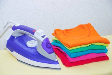 Demir ve bebek kıyafetleri. Ütü tahtasında renkli kıyafetler. Parlak tişörtler. Bir tahta üzerinde ütülenmiş renkli bebek iç çamaşırı. . Ütü tahtası. Bir yığın şey.