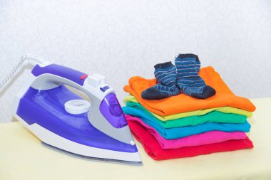 Demir ve bebek kıyafetleri. Ütü tahtasında renkli kıyafetler. Parlak tişörtler. Bir tahta üzerinde ütülenmiş renkli bebek iç çamaşırı. . Ütü tahtası. Bir yığın şey.