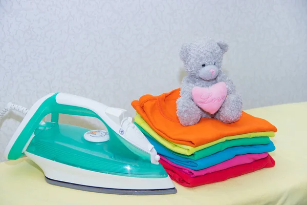 Iron and baby clothes. Colored clothes on an ironing board. Bright t-shirts. Ironed colored baby underwear on a blackboard. . Ironing board. A pile of things.