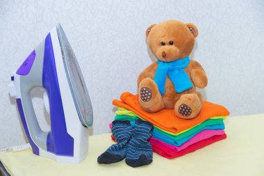 Demir ve bebek kıyafetleri. Ütü tahtasında renkli kıyafetler. Parlak tişörtler. Bir tahta üzerinde ütülenmiş renkli bebek iç çamaşırı. . Ütü tahtası. Bir yığın şey.
