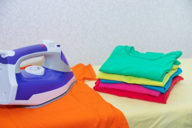 Demir ve bebek kıyafetleri. Ütü tahtasında renkli kıyafetler. Parlak tişörtler. Bir tahta üzerinde ütülenmiş renkli bebek iç çamaşırı. . Ütü tahtası. Bir yığın şey.