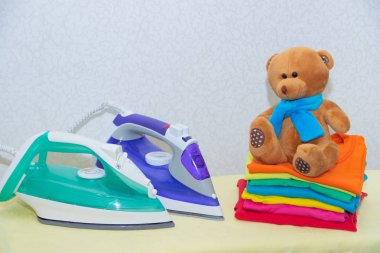 Demir ve bebek kıyafetleri. Ütü tahtasında renkli kıyafetler. Parlak tişörtler. Bir tahta üzerinde ütülenmiş renkli bebek iç çamaşırı. . Ütü tahtası. Bir yığın şey.