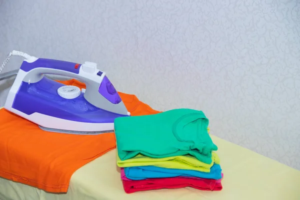 Iron and baby clothes. Colored clothes on an ironing board. Bright t-shirts. Ironed colored baby underwear on a blackboard. . Ironing board. A pile of things.