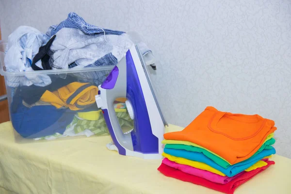 Iron and baby clothes. Colored clothes on an ironing board. Bright t-shirts. Ironed and non-ironed colored children\'s underwear on the board. Ironing board. A pile of things.