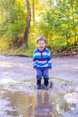 Küçük bir çocuk su birikintisinde zıplıyor. Lastik çizmeli bir çocuk. Mutlu çocukluk. Yağmurdan sonra su birikintileri. Sıcak yaz akşamı.