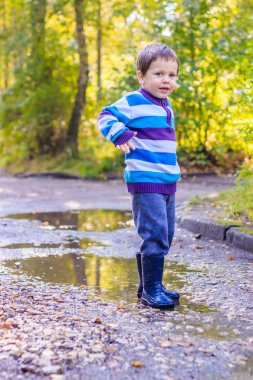 Küçük bir çocuk su birikintisinde zıplıyor. Lastik çizmeli bir çocuk. Mutlu çocukluk. Yağmurdan sonra su birikintileri. Sıcak yaz akşamı.