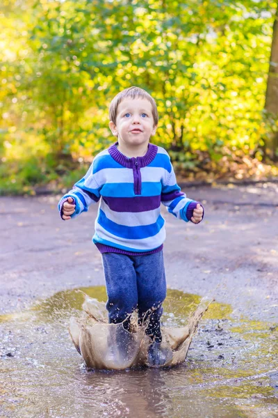 Um rapazinho está a saltar para uma poça. Um rapaz de botas de borracha. Feliz infância. Pudim depois da chuva. Quente noite de verão . — Fotografia de Stock